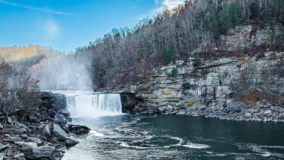 A wide waterfall flows over a rocky cliff into a river surrounded by a forest.